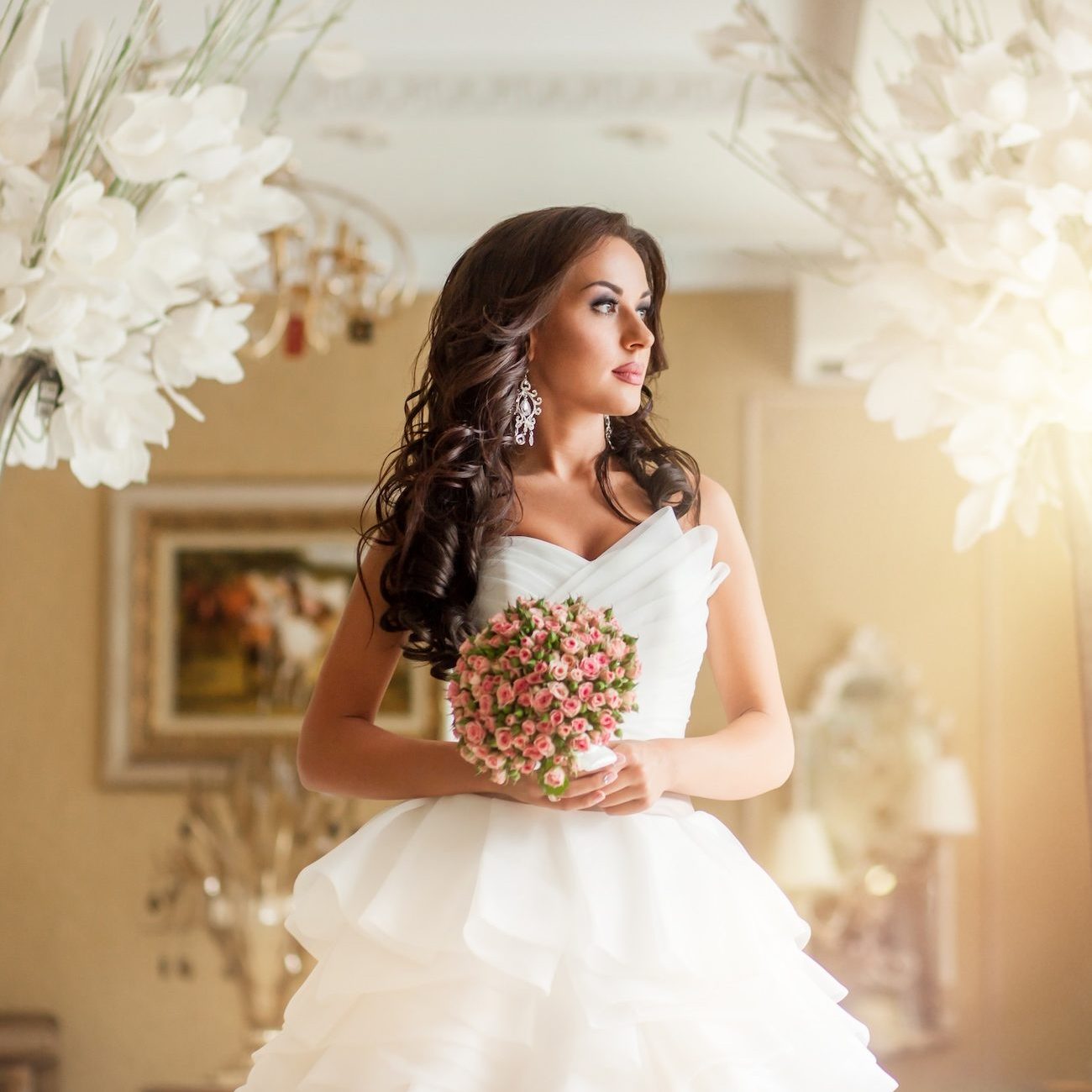 Beauty adult woman bride in white wedding dress in luxury place A woman in a white wedding gown holds a bouquet of flowers, standing between two tall floral arrangements in a decorated room.