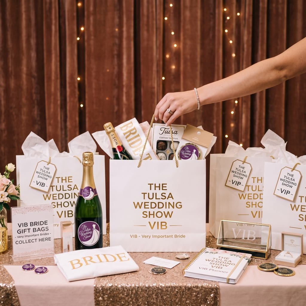 A hand reaches for a gift bag labeled “The Tulsa Wedding Show VIB” among several bags, champagne, and bridal event items on a decorated table with a brown curtain backdrop.