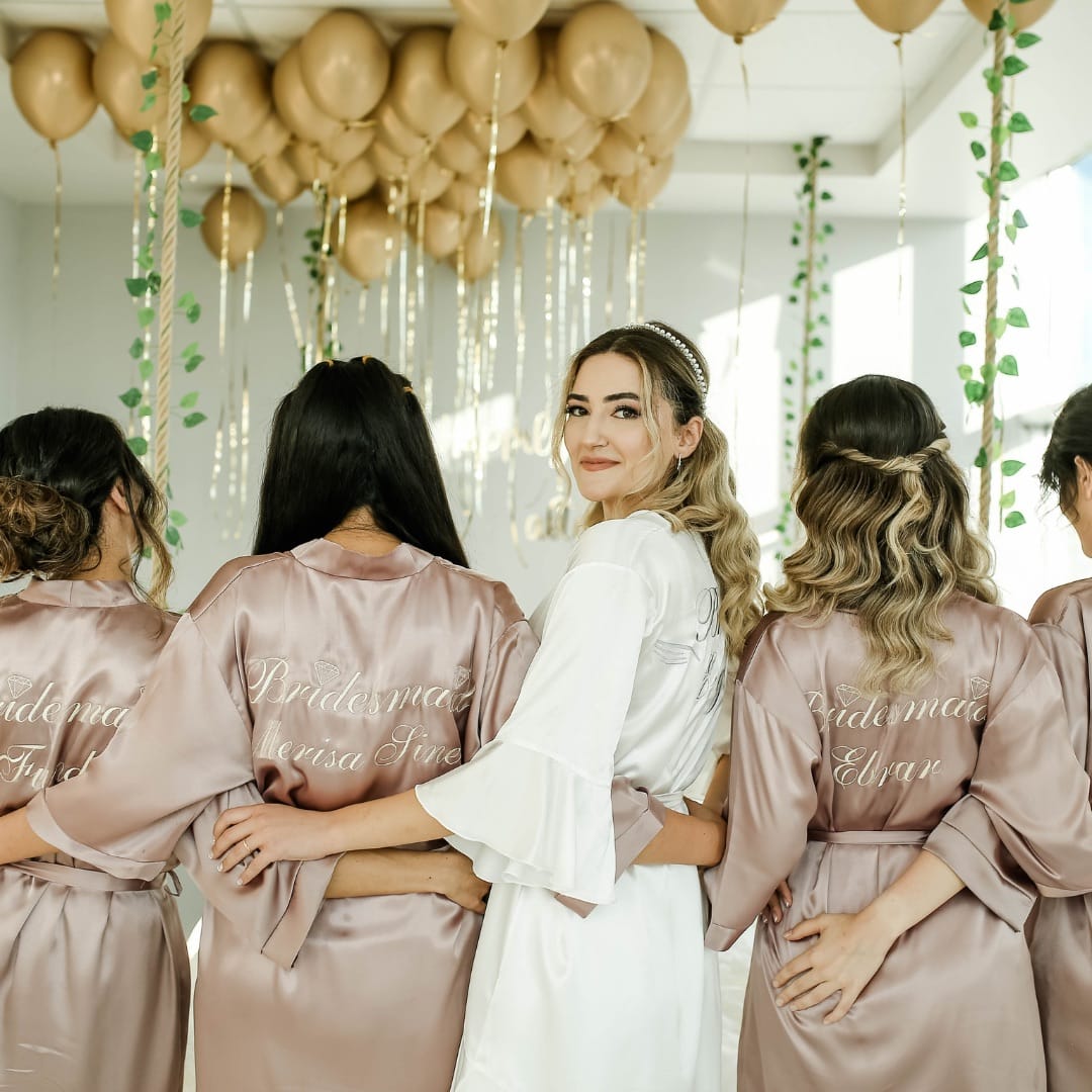 Five women stand together indoors, four in matching bridesmaid robes and one in a white robe, facing away except the woman in white who looks at the camera. Gold balloons and vines decorate the background.